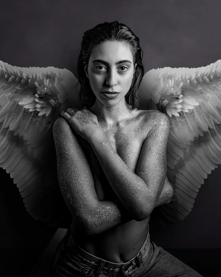 Black-and-white editorial portrait of a model with angel wings and glittered skin